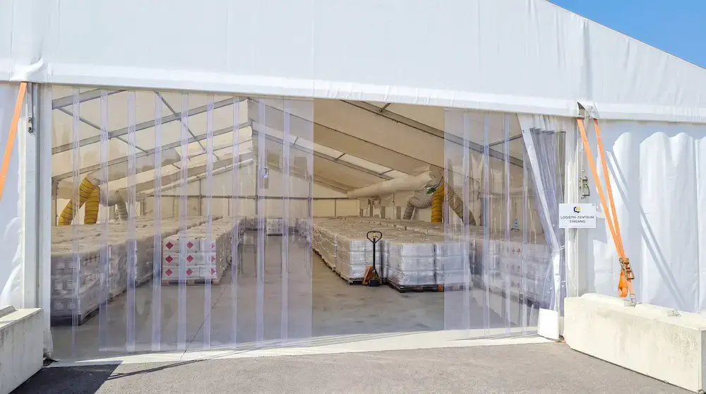 Blick durch einen transparenten Lamellenvorhang in ein großes, weißes Festzelt, das komplett mit palettierten Gefahrgut-Kanistern (Desinfektionsmittel) gefüllt ist; im Hintergrund sind gelbe Luftschläuche der Klimatisierung zu sehen.