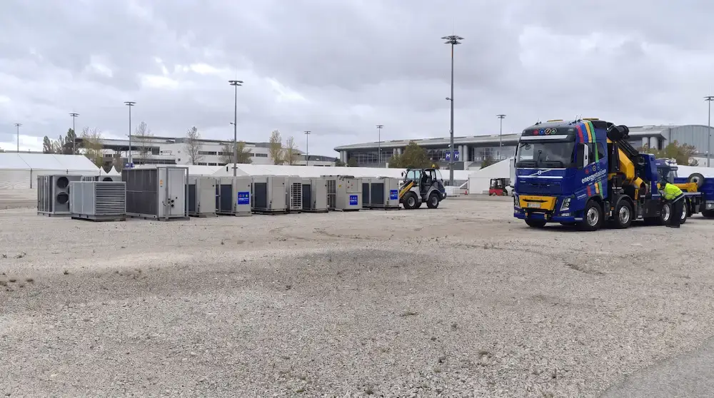 Reihe von mobilen Kältemaschinen und Airhandlern auf Schotterboden vor Messegebäuden, im Hintergrund ein Lkw mit Ladekran und ein Radlader beim Entladen.